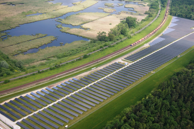 Luftaufnahme eines Solarparks mit Panelen auf einem Feld, umgeben von Bäumen, Gras, Pflanzen und Wasser, mit einem Zug auf einer nahen Bahntrasse.