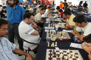 Eine Gruppe von Menschen spielt Schach mit einem Mann in einem blauen Hemd auf der linken Seite, einem Fenster oben rechts und einem Tisch mit einem Glas, Schachbrett, Schachfiguren, einer Coke, Papier, Stift und einer Tischdecke.