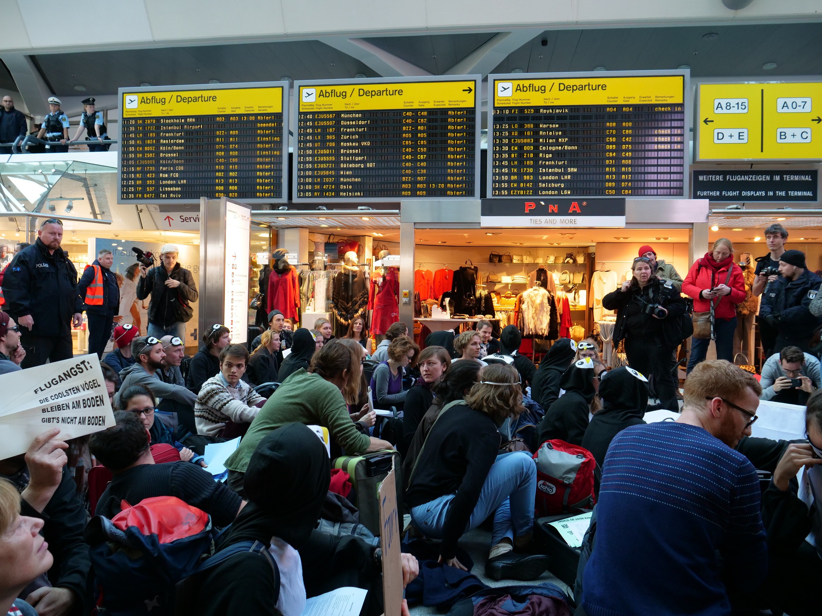 Eine große Gruppe von Menschen sitzt und steht in einem Flughafen während einer Demonstration, wobei einige Taschen und Papiere halten, während Bretter mit Text, Schaufensterpuppen in Kleidern und Deckenleuchten im Hintergrund zu sehen sind.
