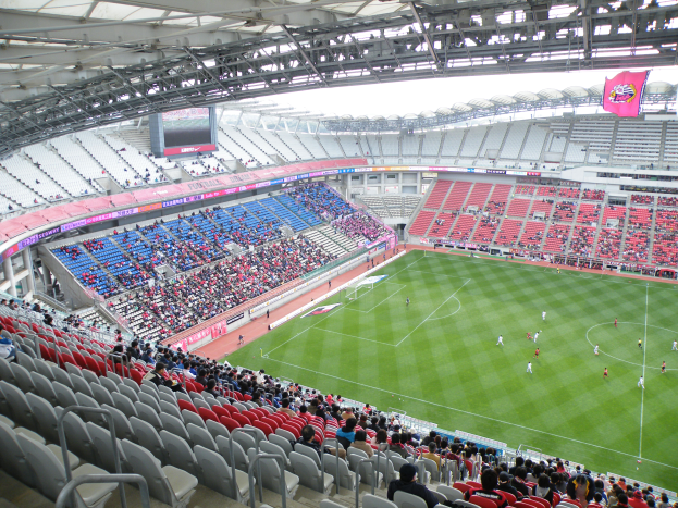 Ein großes Stadion voller Zuschauer, die ein Fußballspiel verfolgen, mit Spielern im Einsatz auf dem Feld und einer Hütte mit Stangen, Bannern und einem Bildschirm oben.