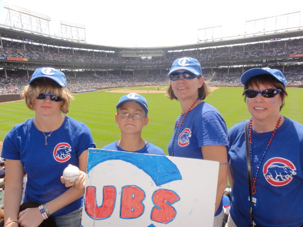 Drei Frauen in Kappen und Schutzbrillen, ein Junge in Brille und Kappe mit einem Ball und einer Tafel, stehend in einem Stadion mit Publikum und Feld dahinter, unter einem Himmel mit einigen Lichtern.