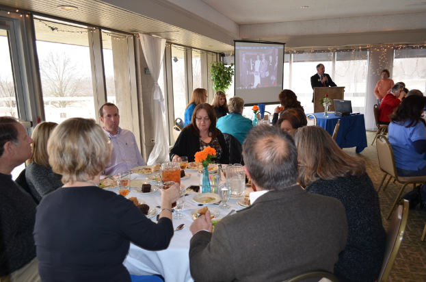 Eine Gruppe von Menschen um einen Tisch versammelt, der mit verschiedenen Speisen gefüllt ist, eine stehende Person, die spricht, ein Projektor, der Inhalte anzeigt, eine Pflanze neben dem Projektor und ein Blick aus dem Fenster mit Vorhängen, Himmel und Bäumen im Hintergrund.