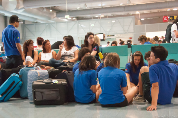 Ein belebter Flughafeninnenraum mit vielen Menschen, die auf Flüge warten, Gepäck links und gläserne Türen mit Richtungsschildern im Hintergrund.