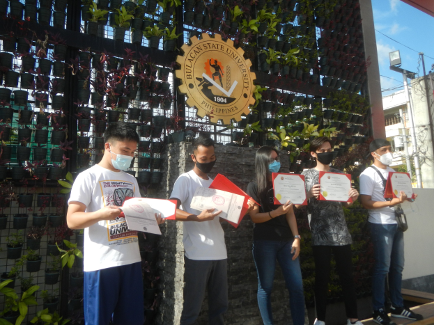 Gruppe von maskierten Menschen mit Urkunden vor einem Universitätsgebäude mit einer Pflanzenwand links und einem Bogen mit Logo rechts.