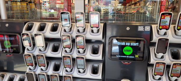 Ein Verkaufsautomat mit mehreren Handys oben drauf, vor einem belebten Hintergrund mit Menschen, Geschäften, Schildern, Lichtern und anderen Gegenständen.