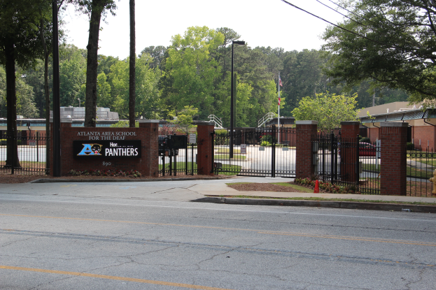 Der Eingang zu einer Schule mit einem Schild an der Seite, umgeben von Bäumen, Straßenlaternen, Gittern, Namensschildern, Kraftfahrzeugen auf der Straße, Absperrgittern, Pflanzen, Gras, Gebäuden und einem klaren blauen Himmel.