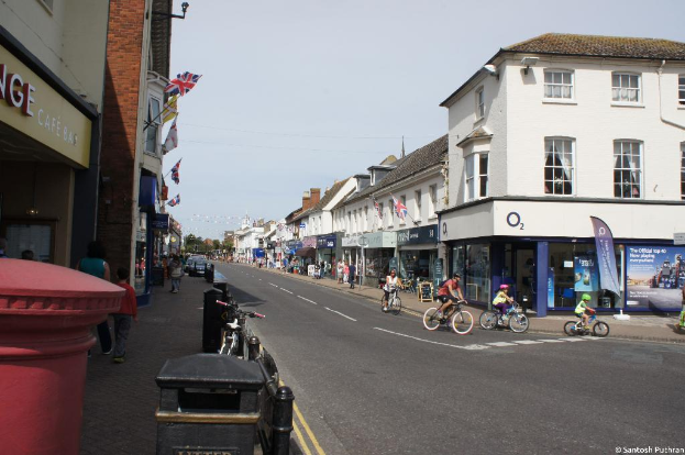 Eine Straße mit Gebäuden auf beiden Seiten, Menschen, die auf der Straße Fahrrad fahren, Fahrzeuge und Fußgänger auf einem Gehweg links daneben, Fahnen an den Gebäuden und ein Banner vor einem Geschäft auf der rechten Seite.