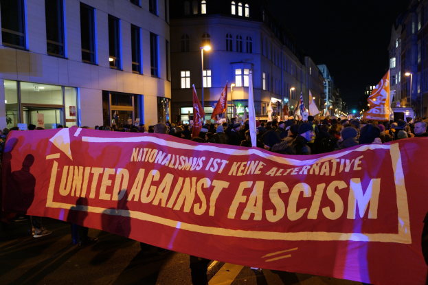 Eine Gruppe von Menschen marschiert die Straße entlang und trägt ein Banner mit der Aufschrift "Einheit gegen Faschismus", während Gebäude, Laternenpfähle und Schilder die Straße säumen und der Himmel sichtbar ist.