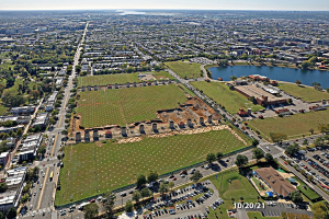 Luftaufnahme einer Stadt mit Gebäuden, Bäumen, Gras, Straßen mit Fahrzeugen, Wasser und Wolken, die einen großen zentralen Bereich zeigen, der als Parkplatz vorgeschlagen ist.