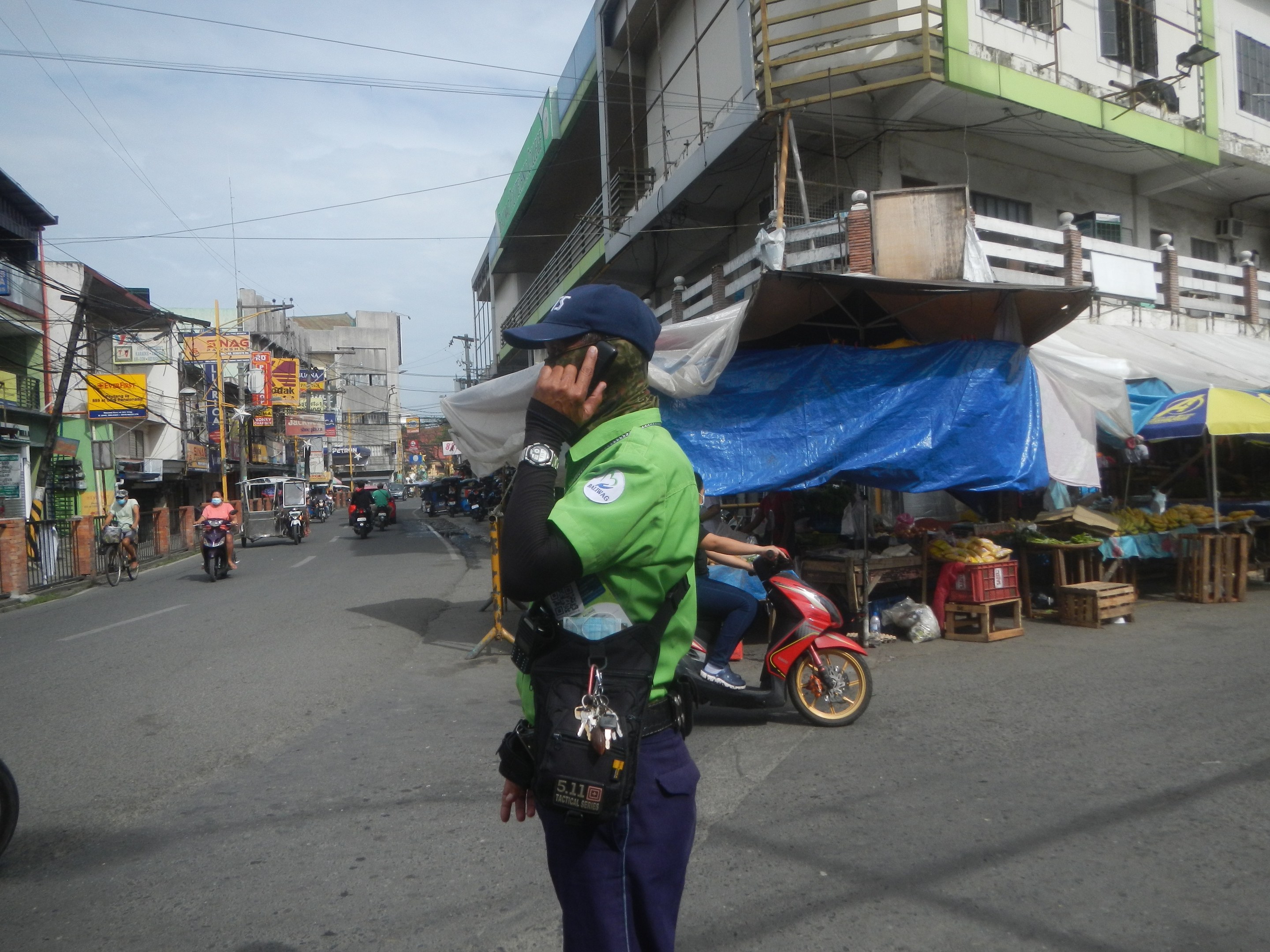 Polizeibeamter in grüner und blauer Uniform mit blauer Mütze, der auf der Straßenseite steht und in ein Handy spricht, mit Fahrzeugen auf der Straße, Marktbuden rechts und einem Gebäude mit Fenstern im bewölkten Himmel im Hintergrund.