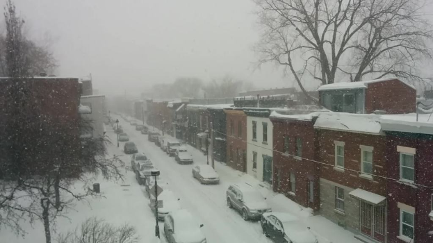 Eine schneebedeckte Straße mit Autos und Gebäuden auf beiden Seiten, mit Bäumen im Hintergrund.