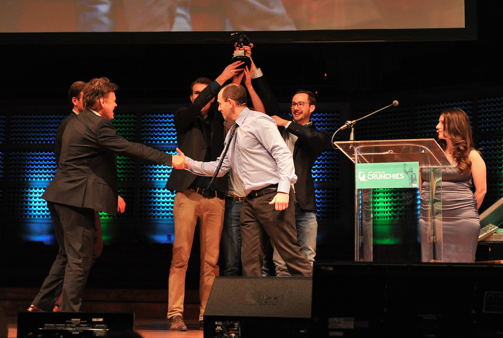 Zwei Männer in schwarzen Mänteln heben einen Preis mit den Händen und lächeln, während ein Mann links die Hand schüttelt und eine Frau rechts steht.