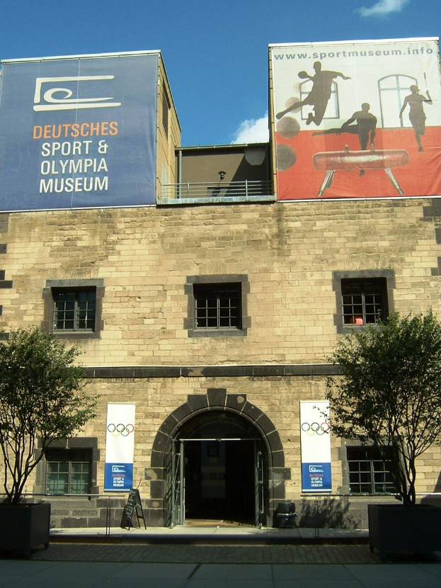 Das Deutsche Sport & Olympiamuseum in Berlin, Deutschland, ein Gebäude mit Fenstern und einer Tür, umgeben von Bäumen und Bannern mit Text und Bildern, unter einem bewölkten Himmel.