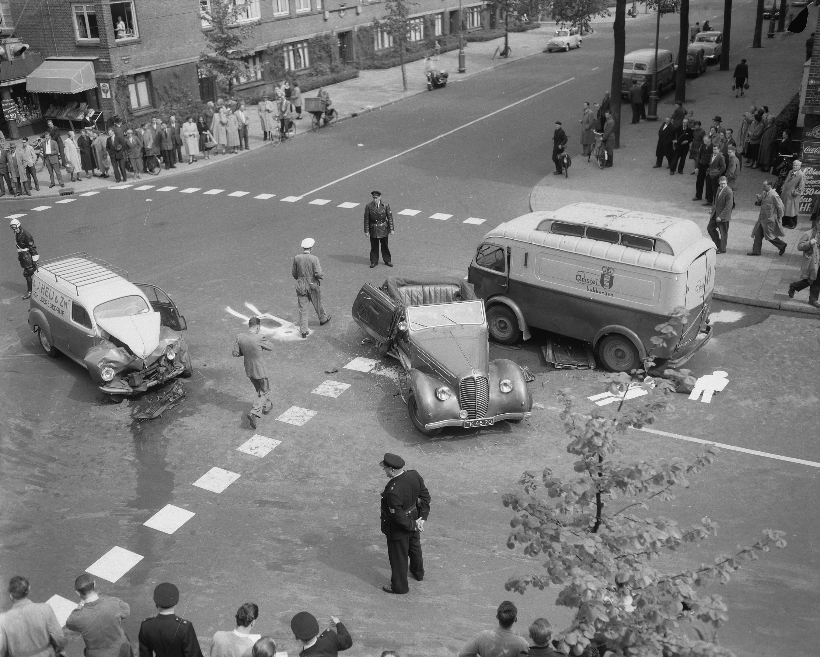 Schwarz-weiß-Szene eines Autounfalls an einer Straßecke mit mehreren Fahrzeugen, darunter ein Van, und einer Gruppe von Menschen in der Nähe, mit Gebäuden, Bäumen, Masten und Texttafeln im Hintergrund.