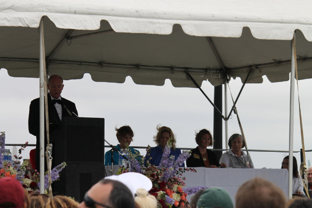Eine Gruppe von Menschen sitzt und steht in der Nähe eines Tisches mit Flaschen, Blumen und einem Mikrofon auf einem Ständer unter einem Zelt mit sichtbaren Polen.