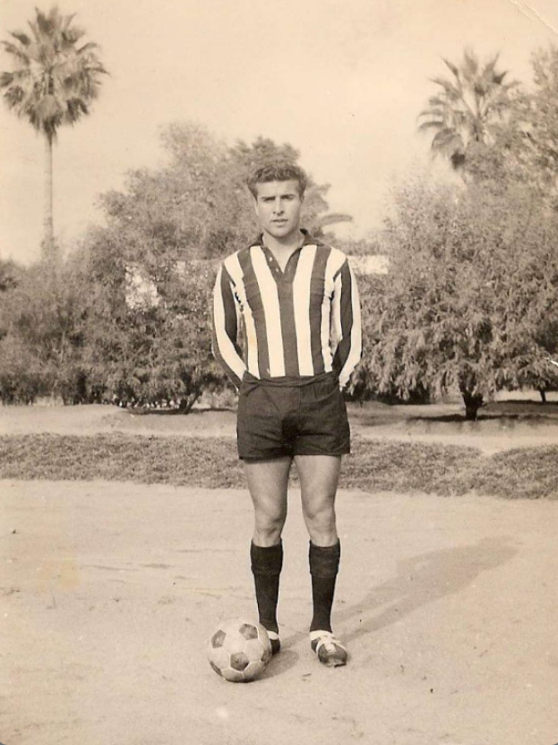 Ein Schwarz-Weiß-Foto eines älteren Mannes, der einen Fußball hält, mit Bäumen und Himmel im Hintergrund.
