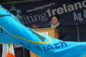 Eine Frau steht an einem Schreibtisch und spricht in ein Mikrofon, mit einem blauen Tuch mit Text davor und einer Wand mit einer Tafel und Werbung im Hintergrund.