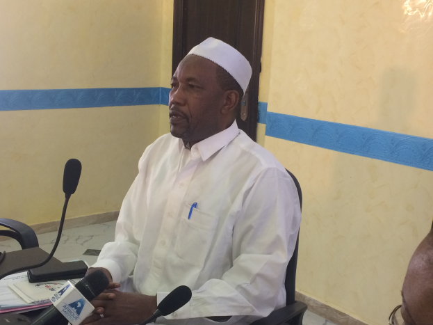 Ein Mann in einem weißen Kleid und Hut sitzt an einem Tisch mit Mikrofonen, spricht, während Papiere und Gegenstände auf dem Tisch liegen, mit einem anderen Menschen mit Brille rechts von ihm in einem Raum mit einer Wand und einer Tür im Hintergrund.