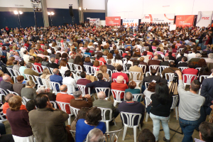 Ein volles Stadion mit vielen sitzenden und stehenden Menschen, eine Bühne mit zwei Rednern am Podium und zahlreichen Bannern und Lichtern.
