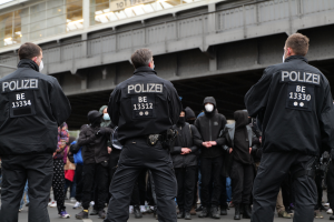 Eine Gruppe von Polizisten in Uniform steht vor einer Menge von Menschen in schwarzen Uniformen und Masken, mit einer Brücke und einem Gebäude im Hintergrund, was auf eine städtische Umgebung hinweist.
