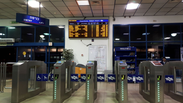 Ein Mann steht vor einer Metalldrehsperre am Flughafen, mit Metalldetektoren, Hinweistafeln, einem Topf mit Pflanze, einer Wandtafel, einer Decke mit Leuchten und einem Bildschirm.
