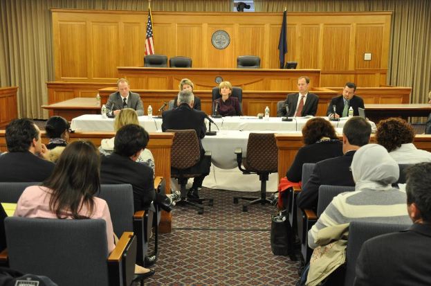 Ein Versammlungsraum mit einer Gruppe von Menschen, die auf Stühlen sitzen, zwei Flaggen anderer Länder, ein oranger Schreibtisch mit Wasserflaschen und einem Mikrofon.
