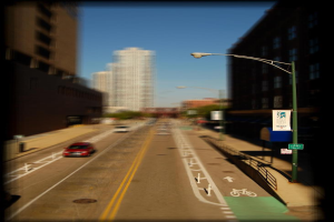 Eine Straßenszene mit Autos auf der Straße, einem Fahrradweg, einer Straßenlaterne mit einem Banner und einem Schild, Gebäuden und Wolkenkratzern im Hintergrund und Fahrbahnmarkierungen.