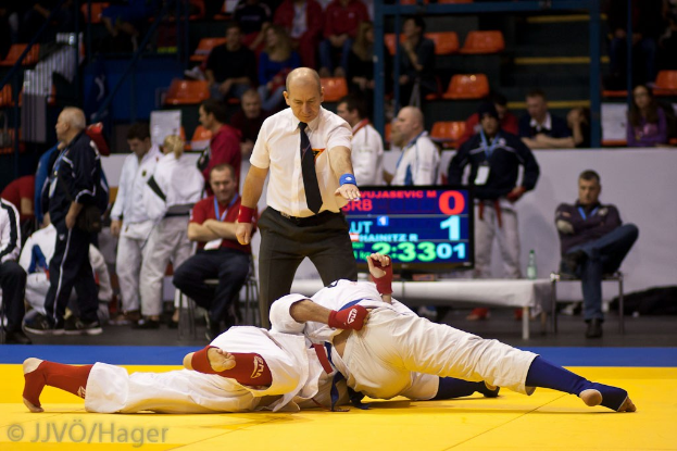 Zwei Judoka im Kampf mit einem Schiedsrichter in der Nähe, in einer Halle mit Zuschauern, Geländern, Treppen und einer Anzeigetafel im Hintergrund.