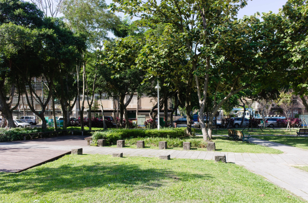 Eine Parkszene mit grünem Gras, Bänken, Pflanzen, Bäumen, Laternenpfählen und Fahrzeugen auf einer Straße, mit Gebäuden und einem klaren blauen Himmel im Hintergrund.