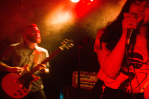 Eine Frau singt in ein Mikrofon auf der Bühne, während ein Mann Gitarre spielt in in Hintergrund unter Deckenlampen.