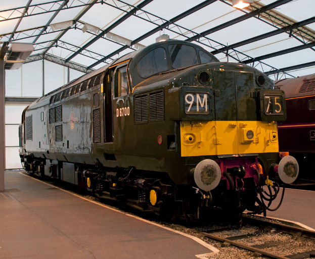 Ein Zug befindet sich auf den Schienen mit einem weiteren Zug neben einem Bahnsteig auf der rechten Seite; der Bahnsteig auf der linken Seite hat einen Mast mit Metallstangen und Lampen daran.