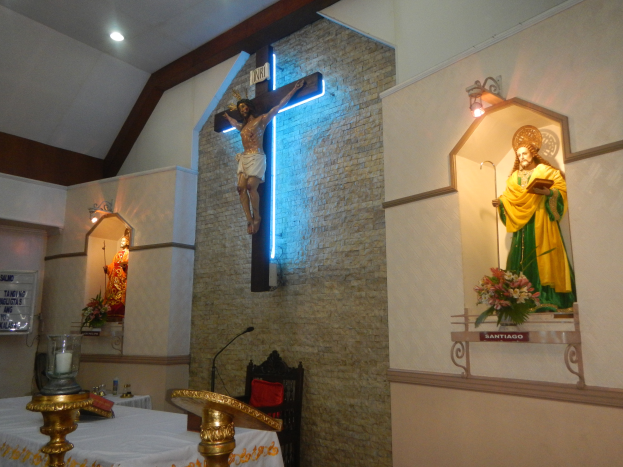Das Innere einer Kirche mit einem Kreuz an der Wand, einer Jesus-Statue am Kreuz, einem Tisch mit einer Decke und verschiedenen Gegenständen, einem Stuhl, einem Mikrofon mit Ständer, einer Tafel mit Text auf der linken Seite, Deckenleuchten und Blumensträußen auf der rechten Seite.