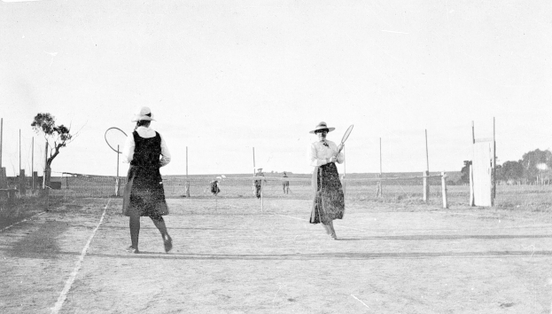 Zwei Frauen spielen Tennis auf einem von Bäumen und Pfosten umgebenen Platz, halten Rackets, in Schwarz-Weiß.