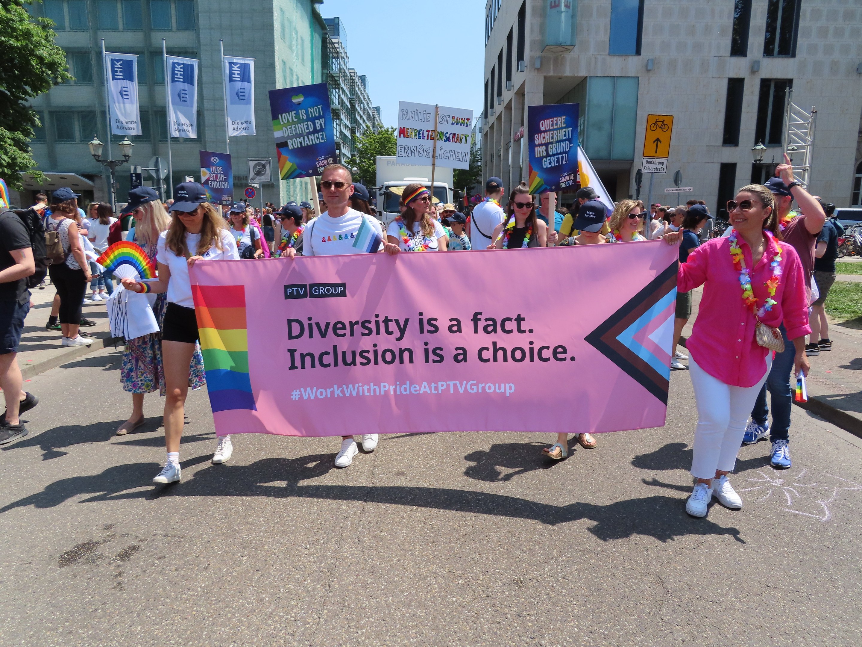 Eine Gruppe von Menschen marschiert auf einer Straße, hält ein Banner mit der Aufschrift "Diversität ist eine Tatsache, Inklusion ist eine Wahl" und trägt teilweise Mützen und Schutzbrillen, während andere zusätzliche Banner halten, vor einem Hintergrund von Gebäuden, Bäumen, Laternen und einem klaren blauen Himmel.