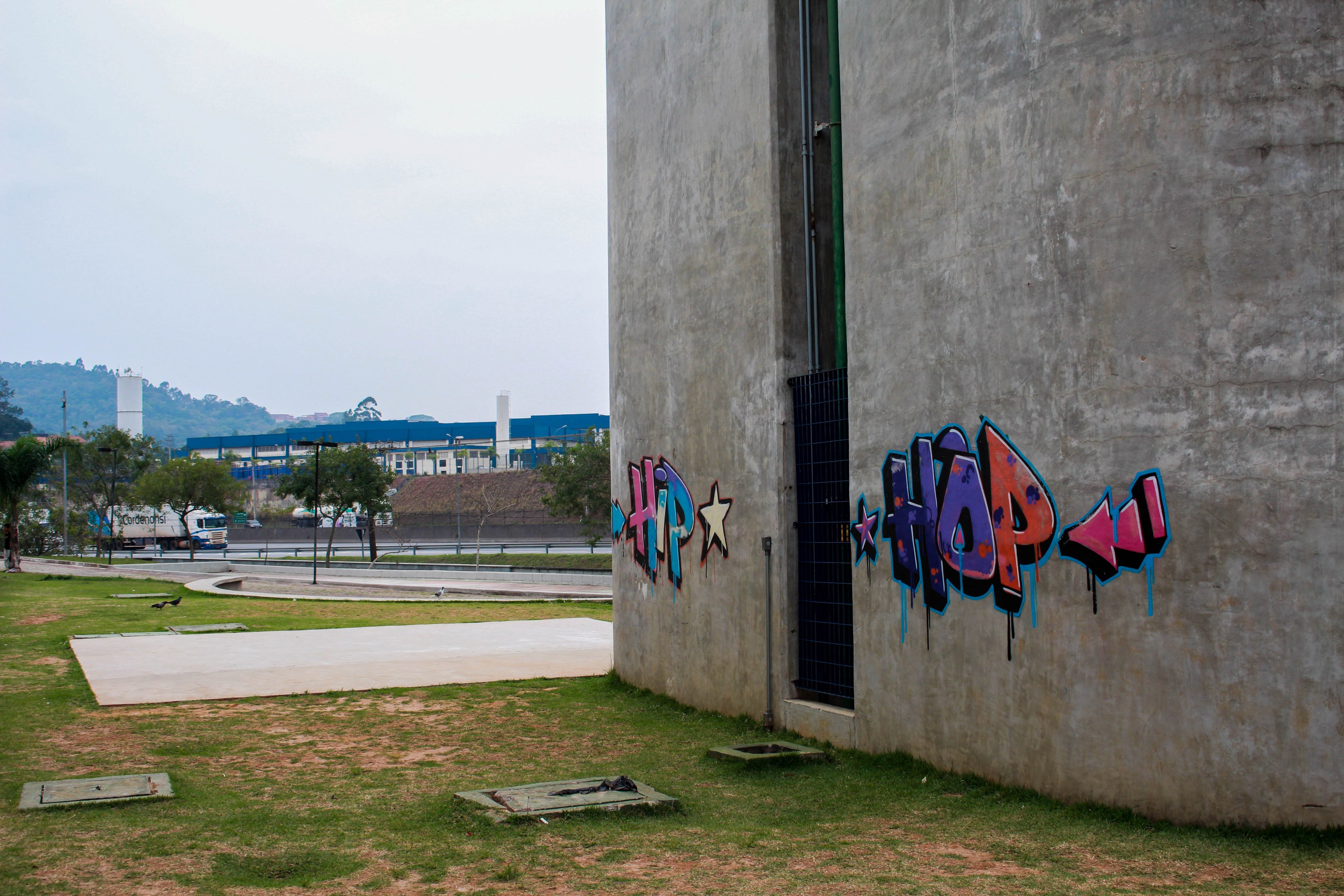 Betongebäude mit Graffiti, umgeben von Gras, Bäumen, Strommasten, Fahrzeugen auf einer Straße, anderen Gebäuden, Hügeln und einem klaren blauen Himmel.
