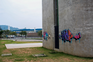 Betongebäude mit Graffiti, umgeben von Gras, Bäumen, Strommasten, Fahrzeugen auf einer Straße, anderen Gebäuden, Hügeln und einem klaren blauen Himmel.