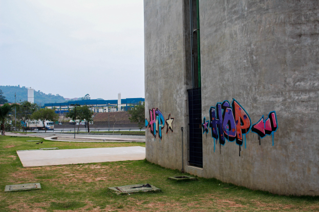 Betongebäude mit Graffiti, umgeben von Gras, Bäumen, Strommasten, Fahrzeugen auf einer Straße, anderen Gebäuden, Hügeln und einem klaren blauen Himmel.
