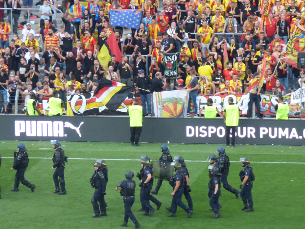 Eine Gruppe von Polizisten in Helmen und mit Waffen in der Hand läuft über ein Fußballfeld, im Hintergrund halten Menschen Fahnen und Banner in der Nähe von Absperrungen.