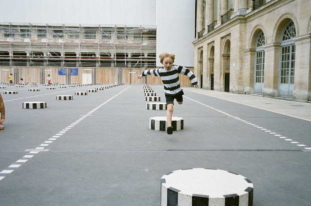 Ein Spielplatz mit vier Kindern, eines läuft auf schwarz-weißen Betonstufen, umgeben von einem Schulgebäude.