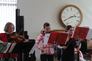 Drei Frauen auf der Bühne mit Geigen spielend, mit Notenpulten und Noten davor, während ein Zuschauer zuhört; eine Uhr ist an der weißen Wand im Hintergrund angebracht, und ein weißer Vorhang ist links zu sehen.