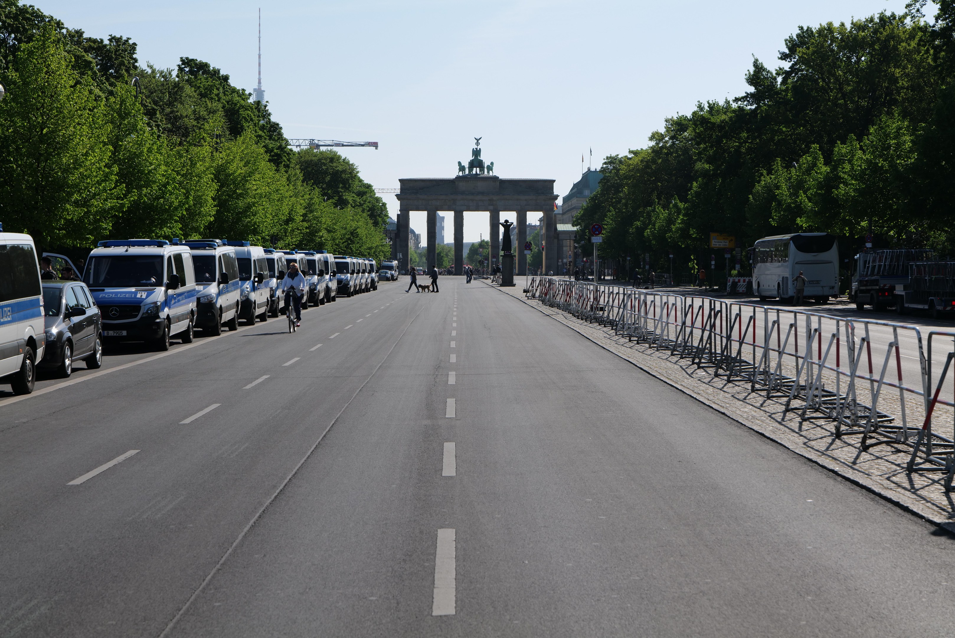 Lange Reihe von Polizeiwagen, die an einer Straße vor dem Brandenburger Tor geparkt sind, mit Menschen auf Fahrrädern und Barrieren auf der Straße, Bäumen an den Seiten und einem Bogen mit Statuen im Hintergrund.