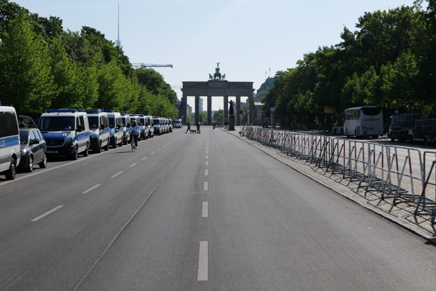 Lange Reihe von Polizeiwagen, die an einer Straße vor dem Brandenburger Tor geparkt sind, mit Menschen auf Fahrrädern und Barrieren auf der Straße, Bäumen an den Seiten und einem Bogen mit Statuen im Hintergrund.