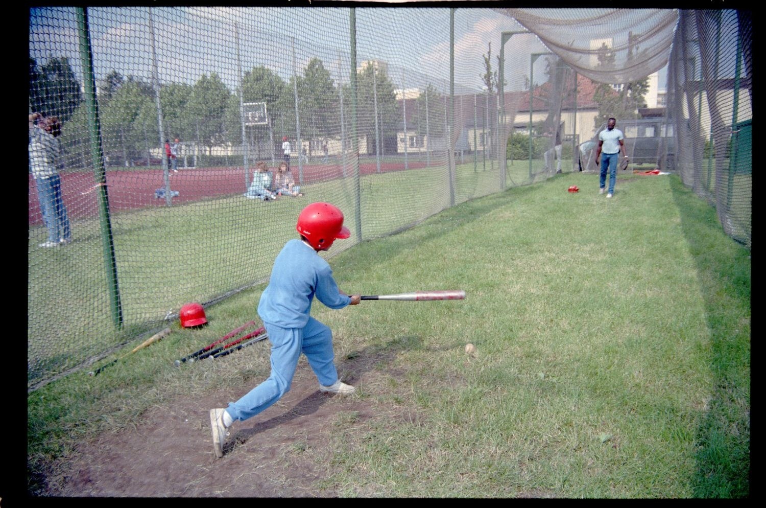 Ein junger Junge in einem blauen Kleid und rotem Helm schwingt einen Baseballschläger auf einem grünen Feld mit verstreuten Schlägern, Bäumen, Gebäuden und einem klaren blauen Himmel im Hintergrund.
