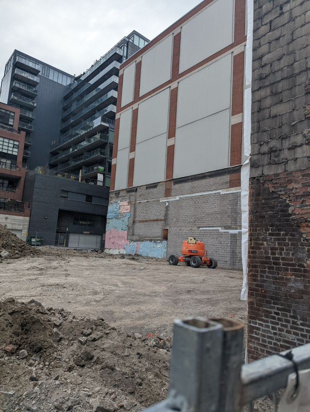 Baustelle mit teilweise abgerissenem Gebäude, einem Fahrzeug, einem Metallzaun und einem bewölkten Himmel.