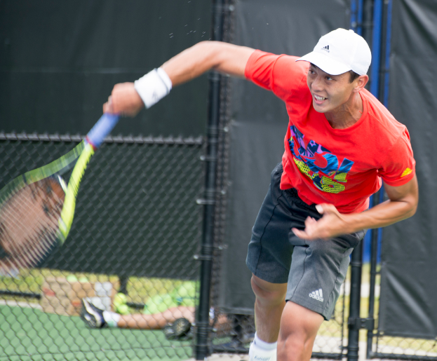 Ein Mann in roter Jacke und weißer Kappe spielt Tennis, hält einen Schläger und hat einen Netz- und durchsichtigen Hintergrund; eine weitere Person sitzt in der Nähe auf dem Boden mit einer Box und Bällen.