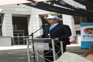Ein Mann in Anzug und Hut spricht in ein Mikrofon an einem Rednerpult, mit einer Person rechts unten, einem Banner mit Text rechts und einem Gebäude mit Gelönder im Hintergrund.