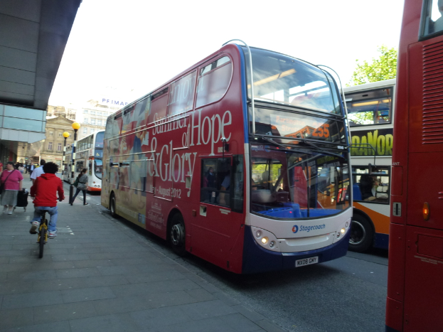 Eine Person fährt Fahrrad auf einem Gehweg mit vielen Menschen, Bussen mit Text darauf, Gebäuden und Laternen im Hintergrund.