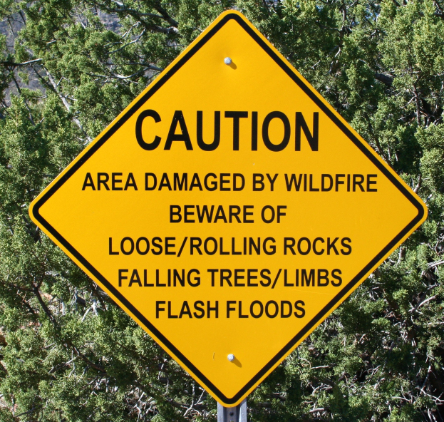 Gelbes Warnschild an einem Pfosten, das vor Wildfire-Schäden, lockeren Steinen, umstürzenden Bäumen und Überschwemmungen warnt.