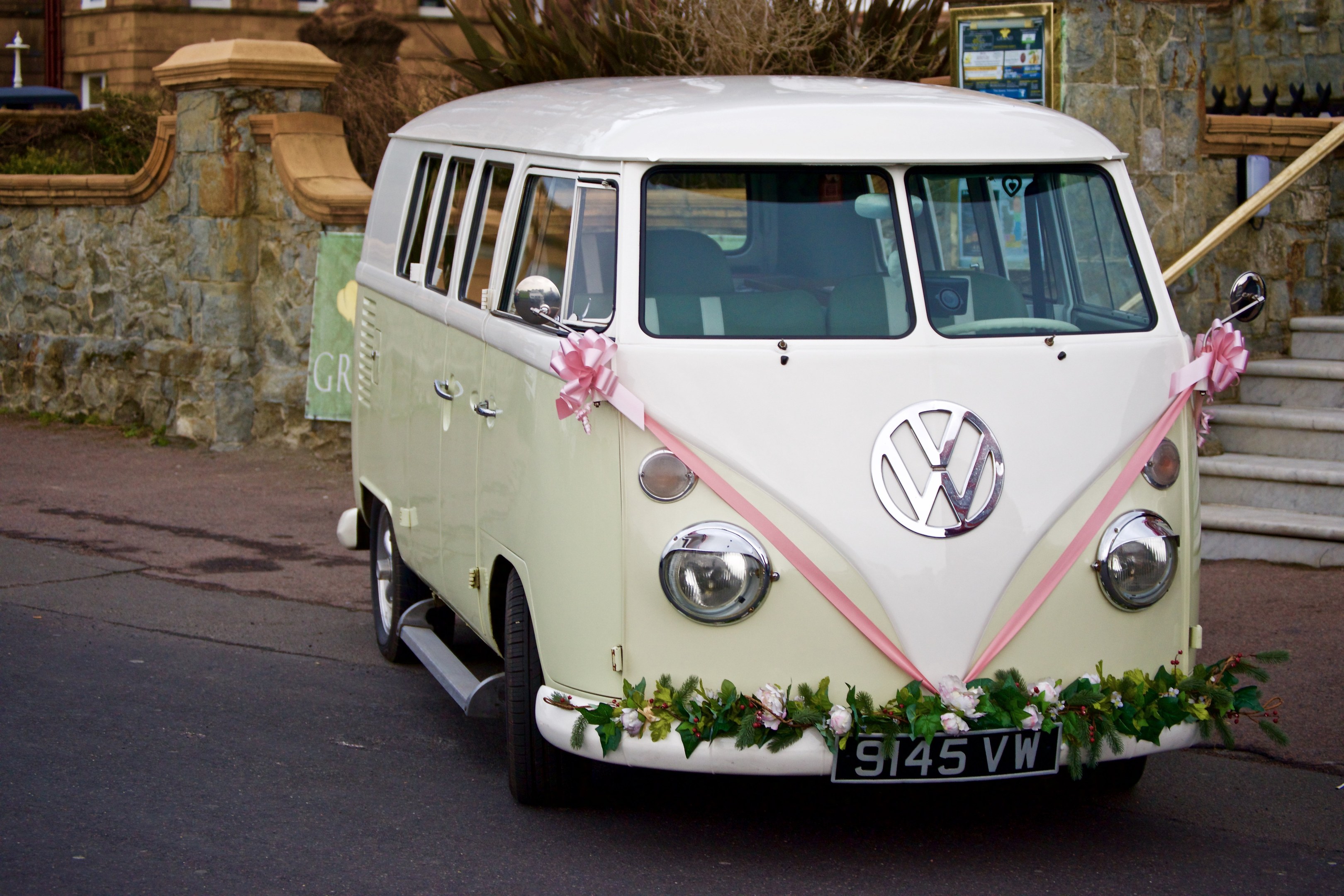 Ein weißer und pinkfarbener VW-Bus mit floralen Verzierungen, der vor einer Steinmauer geparkt ist, neben einem Gebäude mit einer Treppe auf der rechten Seite.