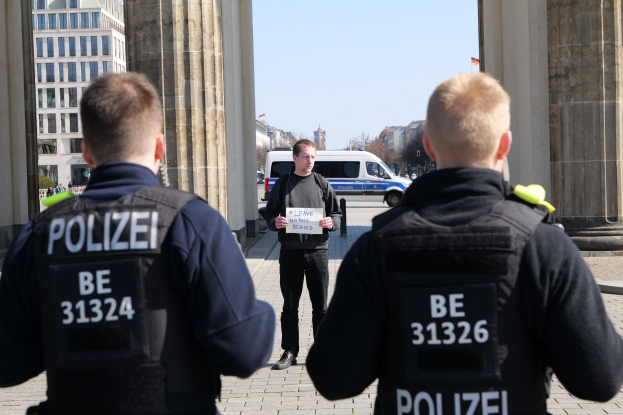 Drei Polizeibeamte vor einem Gebäude mit einem Mann, der ein Papier hält, flankiert von Säulen, mit Fahrzeugen, Menschen, Bäumen, Gebäuden und Himmel im Hintergrund.